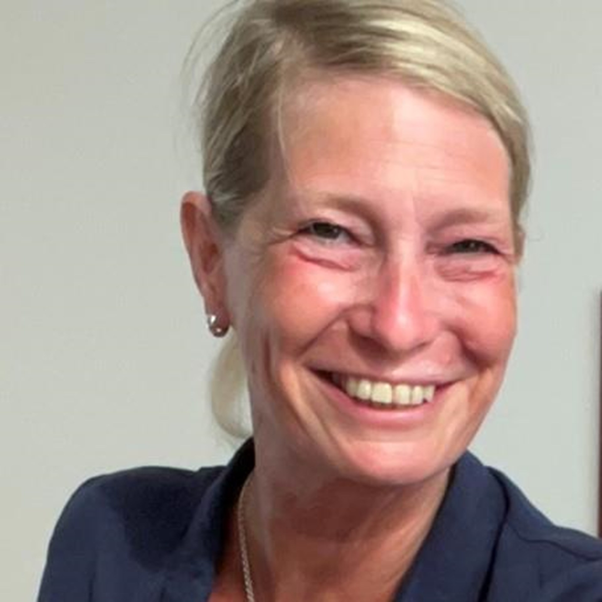 Lächelnde Frau mit blondem Dutt, blauer Bluse und Silberkette vor hellem Hintergrund.