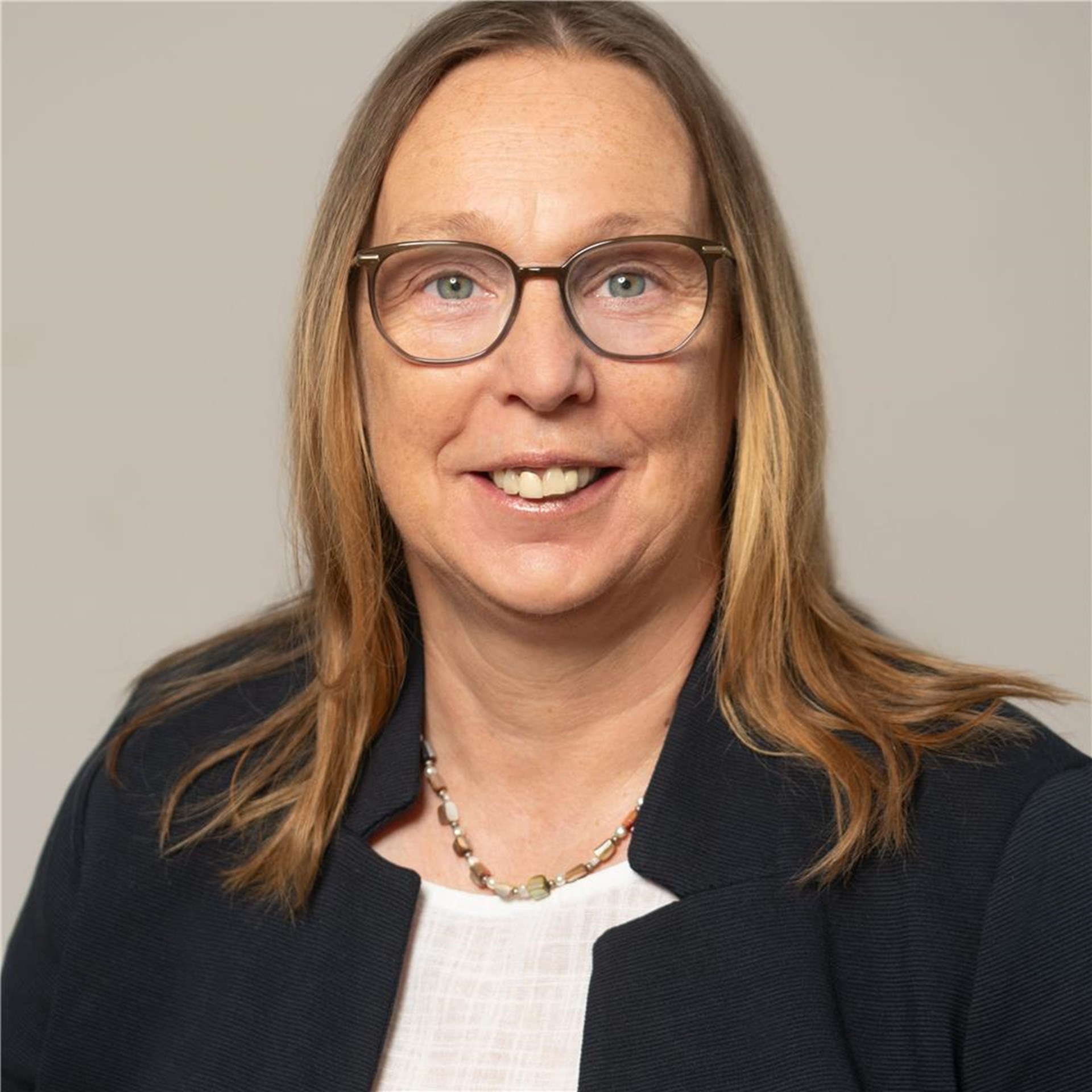 Frau mit schulterlangem, blondem Haar trägt eine schwarze Jacke, weiße Bluse und eine Perlenkette, vor neutralem Hintergrund.